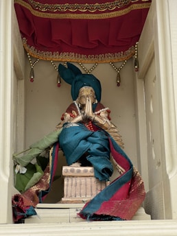 A statue adorned in rich silk fabrics with vibrant teal and maroon colors, sitting on an ornate pedestal. The backdrop features decorative drapery with beadwork trim. The statue is positioned within a niche that frames it, providing a sense of reverence.