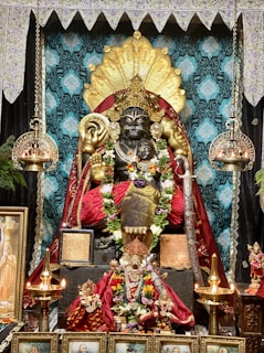 A richly decorated statue depicting a deity adorned with ornate clothing, garlands, and jewelry. The statue is surrounded by an intricately designed altar with brass lamps, floral decorations, and framed religious images. The background features a luxurious tapestry fabric with patterns, and a decorative canopy tops the scene.