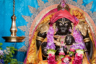 A richly adorned idol with fierce expression and intricate gold embellishments. The statue is decorated with vibrant floral garlands, including pink and purple flowers. A golden headdress and jewelry enhance the idol's presence, and it is set against an ornate backdrop with detailed patterns, featuring a ceremonial lamp on the left.