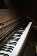 A close-up view of a piano keyboard with black and white keys. The piano has a dark wooden finish, and the lighting creates a moody, intimate atmosphere.