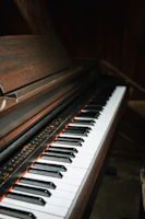 A close-up view of a piano keyboard with black and white keys. The piano has a dark wooden finish, and the lighting creates a moody, intimate atmosphere.