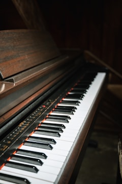 A close-up view of a piano keyboard with black and white keys. The piano has a dark wooden finish, and the lighting creates a moody, intimate atmosphere.