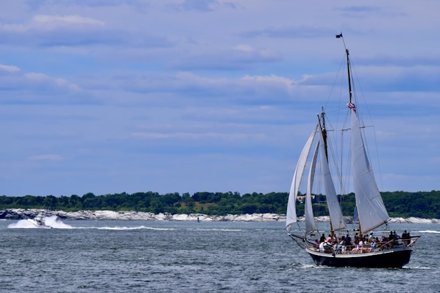 A sailing ship with tall white sails navigates through calm waters. Several people are visible on board, and an American flag is flying from the mast. In the background, a coastline with green trees and rocky formations is under a partly cloudy blue sky.