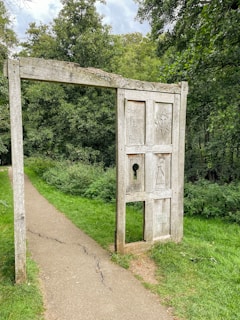 A weathered wooden frame resembling a doorway stands on a grassy path surrounded by dense green foliage. The structure has carvings, including the phrase 'Stories From The Folly' and other engraved symbols. The path is narrow and slightly cracked, leading deeper into the forested area.