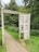 A weathered wooden frame resembling a doorway stands on a grassy path surrounded by dense green foliage. The structure has carvings, including the phrase 'Stories From The Folly' and other engraved symbols. The path is narrow and slightly cracked, leading deeper into the forested area.