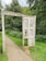 A weathered wooden frame resembling a doorway stands on a grassy path surrounded by dense green foliage. The structure has carvings, including the phrase 'Stories From The Folly' and other engraved symbols. The path is narrow and slightly cracked, leading deeper into the forested area.
