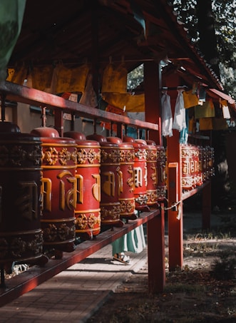 A row of vibrant red and gold prayer wheels, intricately designed with traditional symbols and patterns, is aligned under a wooden structure. The scene is softly lit, casting gentle shadows, and various colorful prayer flags hang above, adding to the spiritual atmosphere.