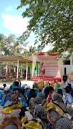 A large group of people are gathered outdoors under the shade of trees. The majority of them are sitting, wearing a variety of colorful outfits and headscarves. In the background, there is a stage decorated with a banner that suggests a community or health-related event. The surroundings include trees and structures, giving a community gathering feel.