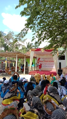 A large group of people are gathered outdoors under the shade of trees. The majority of them are sitting, wearing a variety of colorful outfits and headscarves. In the background, there is a stage decorated with a banner that suggests a community or health-related event. The surroundings include trees and structures, giving a community gathering feel.