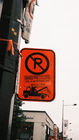 A vividly colored no parking sign is attached to a pole, displaying information about an event called 'Grands Prix Cyclistes' happening on September 10, 2023. Below the no parking symbol, a tow truck icon is visible, indicating towing during the specified event. The background features a blurry view of an urban street with buildings and distant street signs.