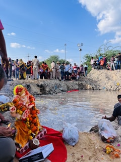 A large group of people gathers near a small water body. In the foreground, a decorated statue of Lord Ganesha is adorned with marigold flowers, resting on a red cloth. Two bags with offerings are placed on the ground. People in the background, some of whom appear to be police officers, are observing the scene.