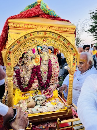 A richly decorated shrine featuring ornate figurines adorned with intricate jewelry and bright floral garlands. Surrounding the shrine, people are engaged in a ritualistic or celebratory activity. An elderly man with yellow markings on his forehead prominently stands beside the shrine, participating in the ceremony. The shrine is golden with red and colorful cloth draped on top.