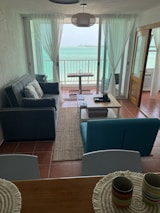 A cozy living room with a view of the ocean through large glass doors. The room has a grey sofa with patterned cushions and a teal armchair. A wooden coffee table is placed on a woven rug. In the background, there is a small table and chair set on a balcony overlooking the sea under a sky that meets the horizon. Sheer white curtains frame the glass doors. A modern, circular ceiling light hangs above.