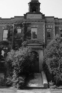 A historic brick building with a classical architectural style, featuring large rectangular windows partially obscured by overgrown vines. Above the entrance, a sign reads 'Academie Salfred'. The building is framed by lush greenery and a path leads to the main door.