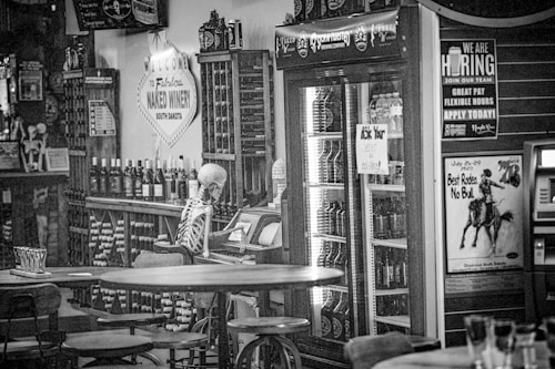 A skeleton sits at a bar counter surrounded by wine bottles and a large refrigerator filled with drinks. The decor includes various signs, one welcoming visitors to a winery in South Dakota, and another advertising employment opportunities. A poster for a rodeo event titled 'Best Rodeo. No Bull.' is visible nearby. The lighting is subdued, creating a somewhat eerie and humorous atmosphere.