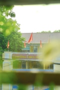 A school entrance with red flags is surrounded by lush greenery, captured from behind blurred foreground foliage. The building has a sloped roof and features a sign with text in a different language.