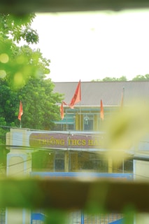 A school entrance with red flags is surrounded by lush greenery, captured from behind blurred foreground foliage. The building has a sloped roof and features a sign with text in a different language.