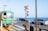 A green tram is passing by a railway crossing near a road with the ocean in the background. Several cars are waiting at the crossing, which is signaled by a traffic light and crossing gate. The clear blue sky and bright sunlight suggest a serene coastal environment.
