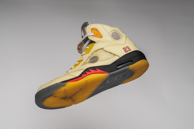 A beige high-top sneaker with a black sole, red and orange accents, and the number 23 printed on the side. The shoe features a Nike logo on the sole and has visible mesh and plastic cutouts throughout its design.