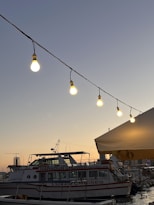 A serene harbor scene at dusk featuring a string of illuminated light bulbs suspended over a cruise ship. The sky displays a gradient from deep blue to a gentle orange near the horizon, reflecting a peaceful evening. A boat labeled 'Harbour Cruises' is docked, accompanied by other vessels in the background. An open canopy provides a partial view to the right.