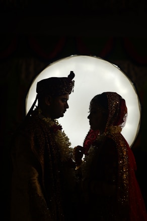 A silhouette of a couple dressed in traditional attire, standing closely with a bright circular light in the background, creating a dramatic contrast.