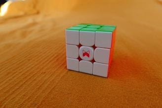 A Rubik's Cube with white, green, and red faces is placed on an orange surface. The cube appears solved from the visible sides, and the background is uniformly textured.