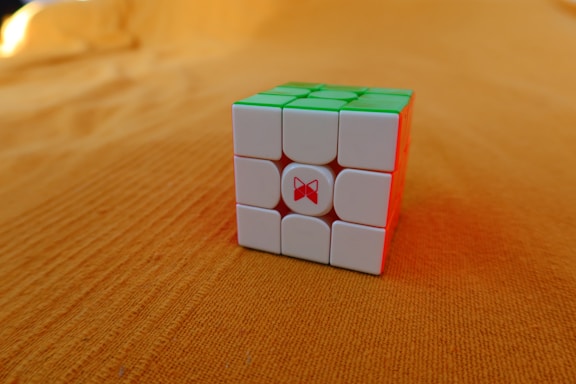 A Rubik's Cube with white, green, and red faces is placed on an orange surface. The cube appears solved from the visible sides, and the background is uniformly textured.