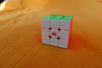 A Rubik's Cube with white, green, and red faces is placed on an orange surface. The cube appears solved from the visible sides, and the background is uniformly textured.