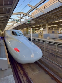 A sleek, modern high-speed train is stationed at a platform under a sheltered area with beams and overhead wires. The train has a streamlined design, with a prominent nose and red lights at the front. The station appears clean and orderly, with various signs and equipment in the background.