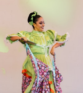A woman is performing a traditional dance, dressed in a vibrant green dress with intricate white lace trim. Her skirt features floral patterns with bright colors such as pink and orange. Her hair is styled with ribbons and flowers matching the dress.