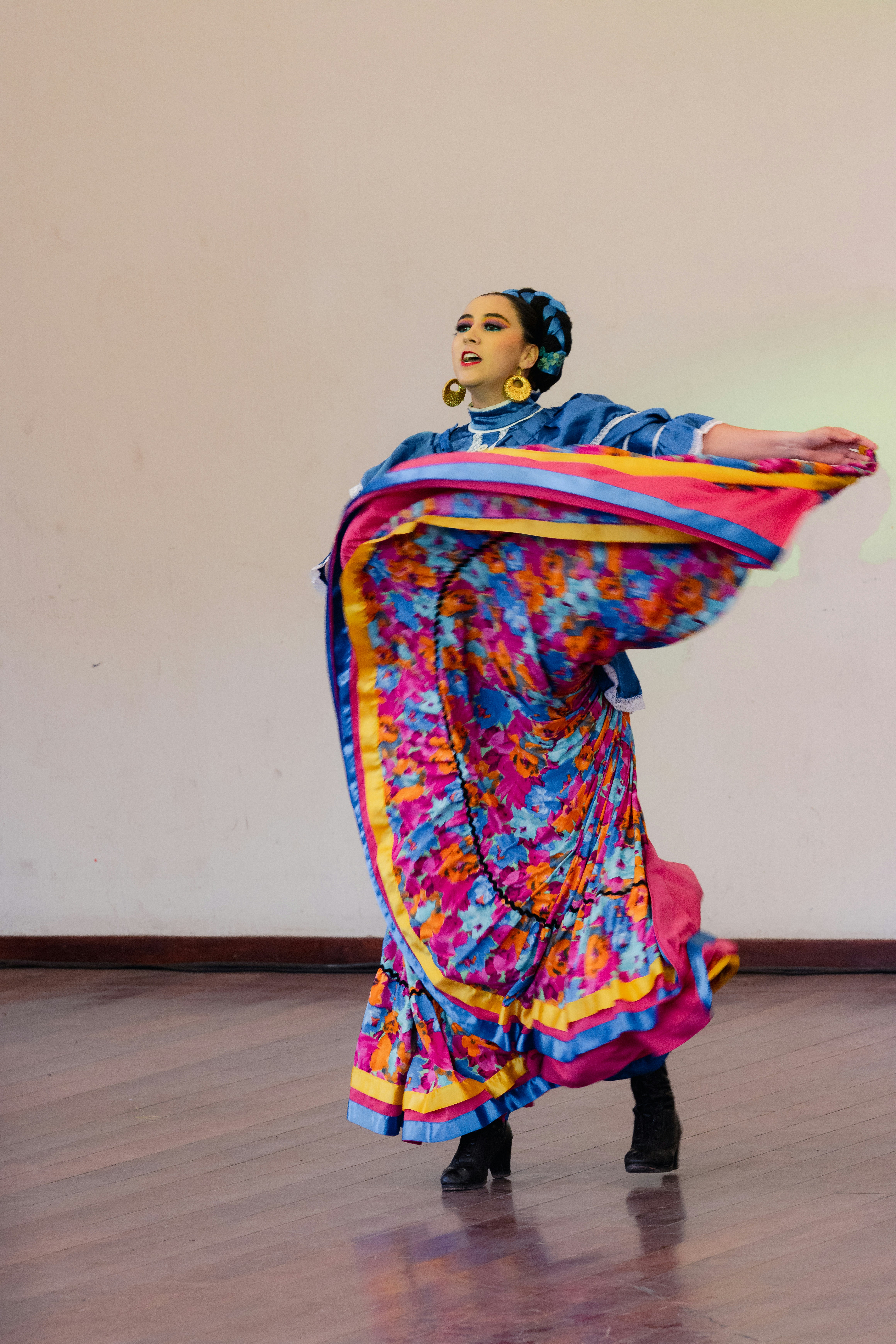 A dancer wearing a colorful traditional dress is gracefully performing on a wooden floor. The outfit features vibrant patterns and bold colors, including blue, pink, yellow, and floral designs. The dancer wears large gold hoop earrings, and her hair is styled neatly with an accessory. The movement of the dance causes the skirt to flare outwards in a dynamic motion.