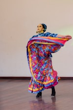 A dancer wearing a colorful traditional dress is gracefully performing on a wooden floor. The outfit features vibrant patterns and bold colors, including blue, pink, yellow, and floral designs. The dancer wears large gold hoop earrings, and her hair is styled neatly with an accessory. The movement of the dance causes the skirt to flare outwards in a dynamic motion.