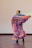 A dancer wearing a colorful traditional dress is gracefully performing on a wooden floor. The outfit features vibrant patterns and bold colors, including blue, pink, yellow, and floral designs. The dancer wears large gold hoop earrings, and her hair is styled neatly with an accessory. The movement of the dance causes the skirt to flare outwards in a dynamic motion.