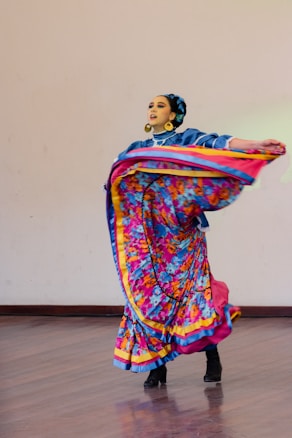 A dancer wearing a colorful traditional dress is gracefully performing on a wooden floor. The outfit features vibrant patterns and bold colors, including blue, pink, yellow, and floral designs. The dancer wears large gold hoop earrings, and her hair is styled neatly with an accessory. The movement of the dance causes the skirt to flare outwards in a dynamic motion.