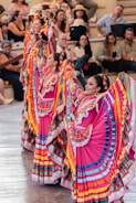 Colorfully dressed dancers, wearing traditional Mexican attire with vibrant skirts and braided hair, perform in front of an audience seated in an amphitheater setting. The performers' dresses feature bright colors like pink, blue, yellow, and green, with intricate lace detailing. Spectators appear engaged, enjoying the lively cultural performance.