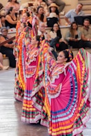 Colorfully dressed dancers, wearing traditional Mexican attire with vibrant skirts and braided hair, perform in front of an audience seated in an amphitheater setting. The performers' dresses feature bright colors like pink, blue, yellow, and green, with intricate lace detailing. Spectators appear engaged, enjoying the lively cultural performance.