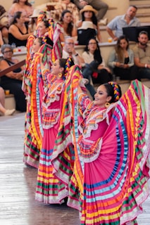 Colorfully dressed dancers, wearing traditional Mexican attire with vibrant skirts and braided hair, perform in front of an audience seated in an amphitheater setting. The performers' dresses feature bright colors like pink, blue, yellow, and green, with intricate lace detailing. Spectators appear engaged, enjoying the lively cultural performance.