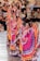 Colorfully dressed dancers, wearing traditional Mexican attire with vibrant skirts and braided hair, perform in front of an audience seated in an amphitheater setting. The performers' dresses feature bright colors like pink, blue, yellow, and green, with intricate lace detailing. Spectators appear engaged, enjoying the lively cultural performance.