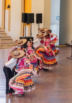 A group of dancers in elaborate, colorful traditional costumes are mid-performance. The women wear vibrant, multi-colored dresses with large skirts, while the men are in white shirts and traditional hats. The setting appears to be an indoor space with wooden flooring and a small set of steps in the background.