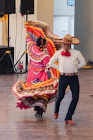 A couple performing a traditional Mexican dance, dressed in vibrant attire. The woman's dress is adorned with multicolored stripes and ruffles, while the man wears a white shirt, red tie, and a large sombrero. The background shows a simple interior setting with speakers.