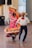 A couple performing a traditional Mexican dance, dressed in vibrant attire. The woman's dress is adorned with multicolored stripes and ruffles, while the man wears a white shirt, red tie, and a large sombrero. The background shows a simple interior setting with speakers.