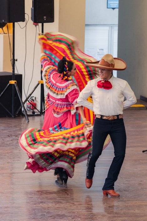 A couple performing a traditional Mexican dance, dressed in vibrant attire. The woman's dress is adorned with multicolored stripes and ruffles, while the man wears a white shirt, red tie, and a large sombrero. The background shows a simple interior setting with speakers.
