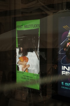 A poster with the text 'FEIST MULTITUDES' is displayed in a glass case. It features an image of a person wearing a white outfit and a brown hat, holding something. The poster is highlighted with a bright green background and contains details about a tour scheduled for September 21, 2023, at the Coliseu de Lisboa. The reflection on the glass suggests an indoor setting.