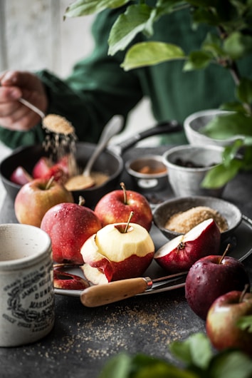 A rustic kitchen setting with several apples, both whole and peeled, placed on a dark countertop. Various small bowls contain ingredients like brown sugar, and a hand is sprinkling sugar over the apples. A knife is placed near the apples, and a mug with vintage writing is visible. A leafy green plant adds a touch of nature to the scene.