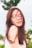 A person with long brown hair and glasses appears content with a serene expression. Their eyes are closed, suggesting a moment of calmness and peace. The background is filled with blurred greenery, giving a sense of being outdoors on a bright day.