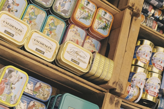 A collection of variously colored tin cans organized on wooden shelves. The tins have different illustrations and labels, each depicting scenes or advertising the contents. The arrangement is dense, and the tins are stacked on top of each other in neatly arranged rows.