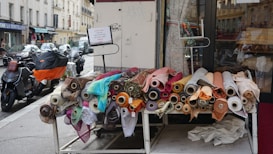 A display of various fabric rolls with different colors and patterns is arranged on a table outside of a shop. The shop's glass door is partially visible, and the surrounding street has parked motorcycles and cars.