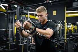 A man is working out in a gym, focused on using a cable machine. He wears a black t-shirt, earphones, and a smartwatch. The gym has various exercise equipment and machinery visible in the background with black and yellow walls.