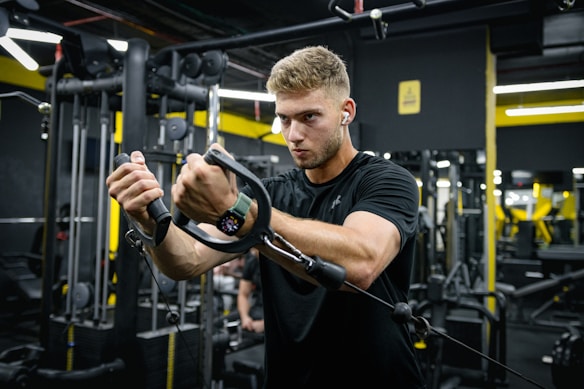 A man is working out in a gym, focused on using a cable machine. He wears a black t-shirt, earphones, and a smartwatch. The gym has various exercise equipment and machinery visible in the background with black and yellow walls.
