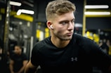 A focused man with short hair wearing black sportswear and wireless earphones stands in a gym. The background features gym equipment and blurred figures, with yellow and black tones dominating the setting.